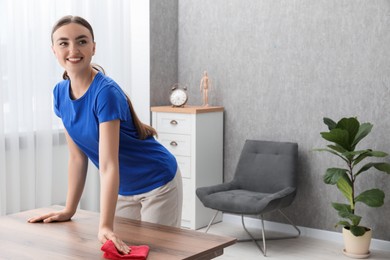 Beautiful young woman cleaning wooden table with rag at home