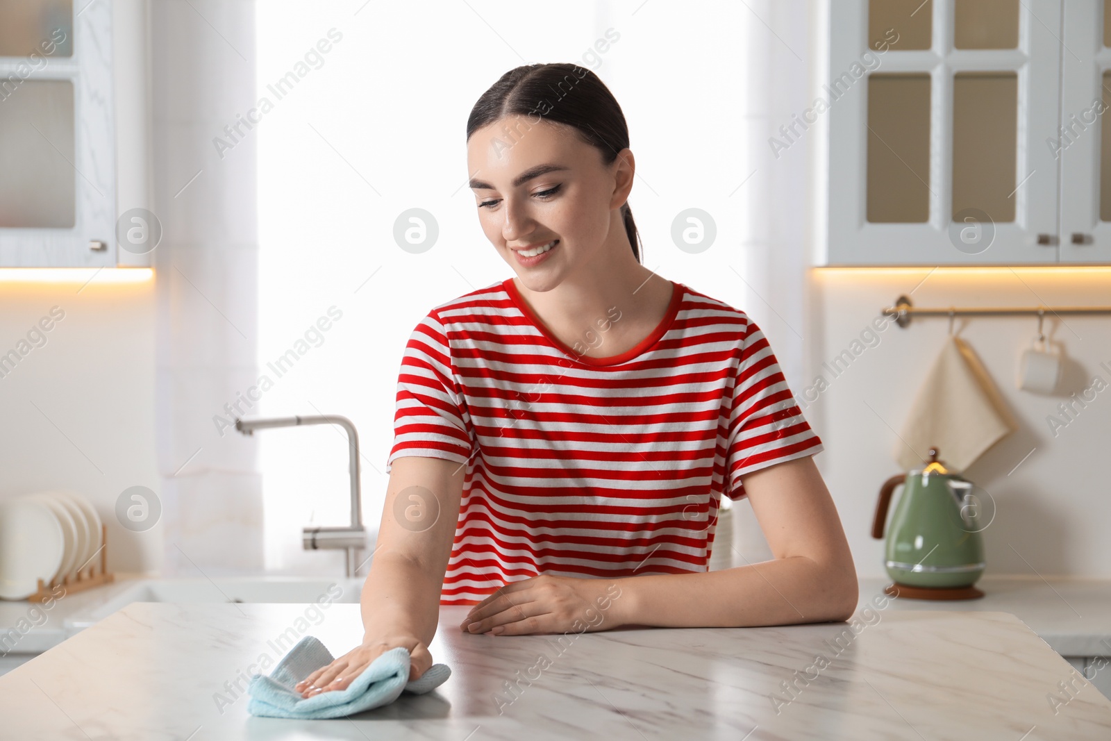 Photo of Beautiful young woman wiping white marble table with rag in kitchen