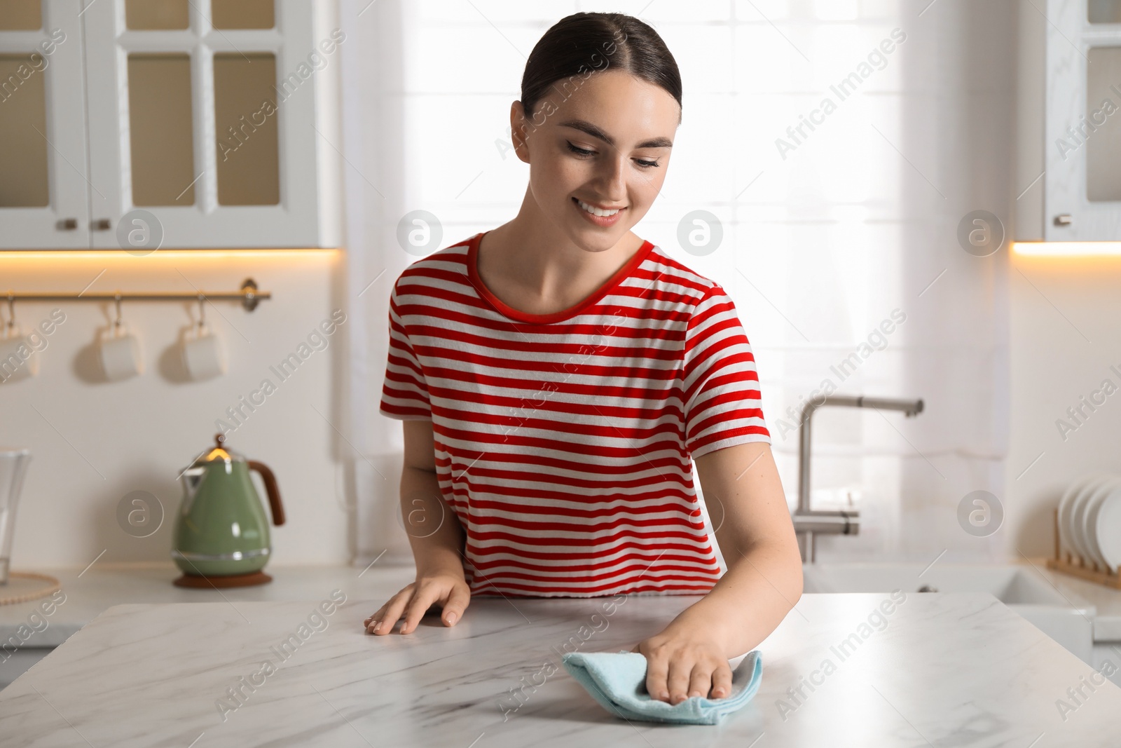 Photo of Beautiful young woman wiping white marble table with rag in kitchen