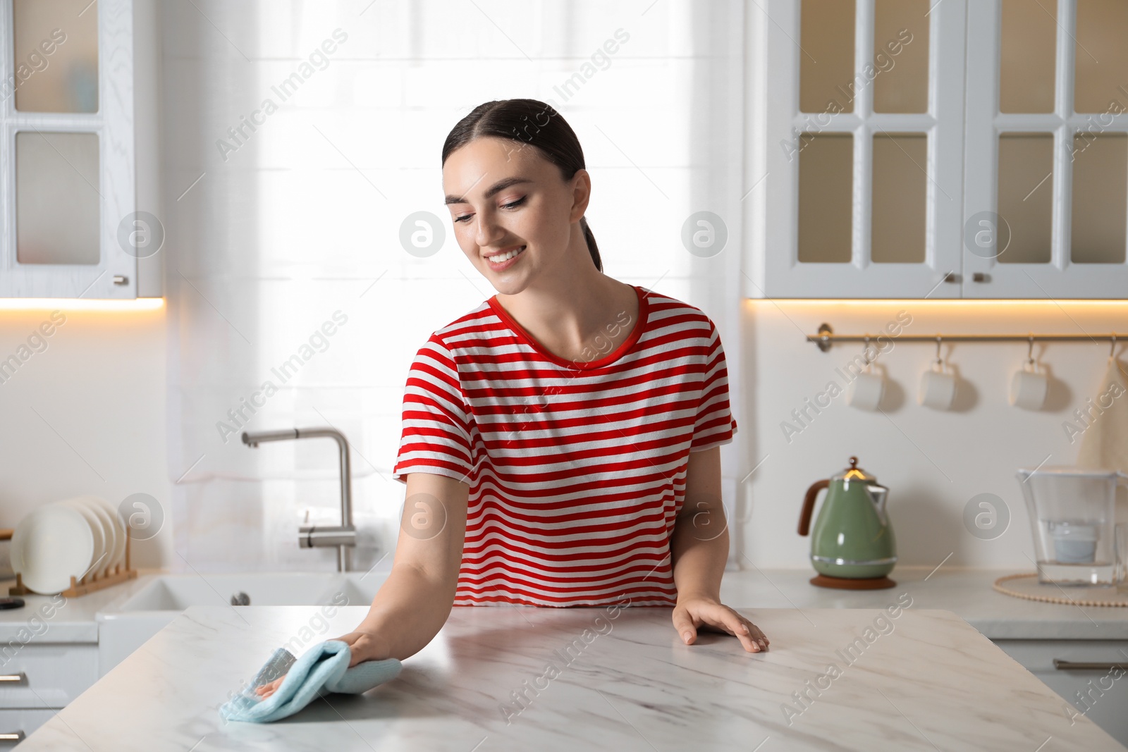 Photo of Beautiful young woman wiping white marble table with rag in kitchen