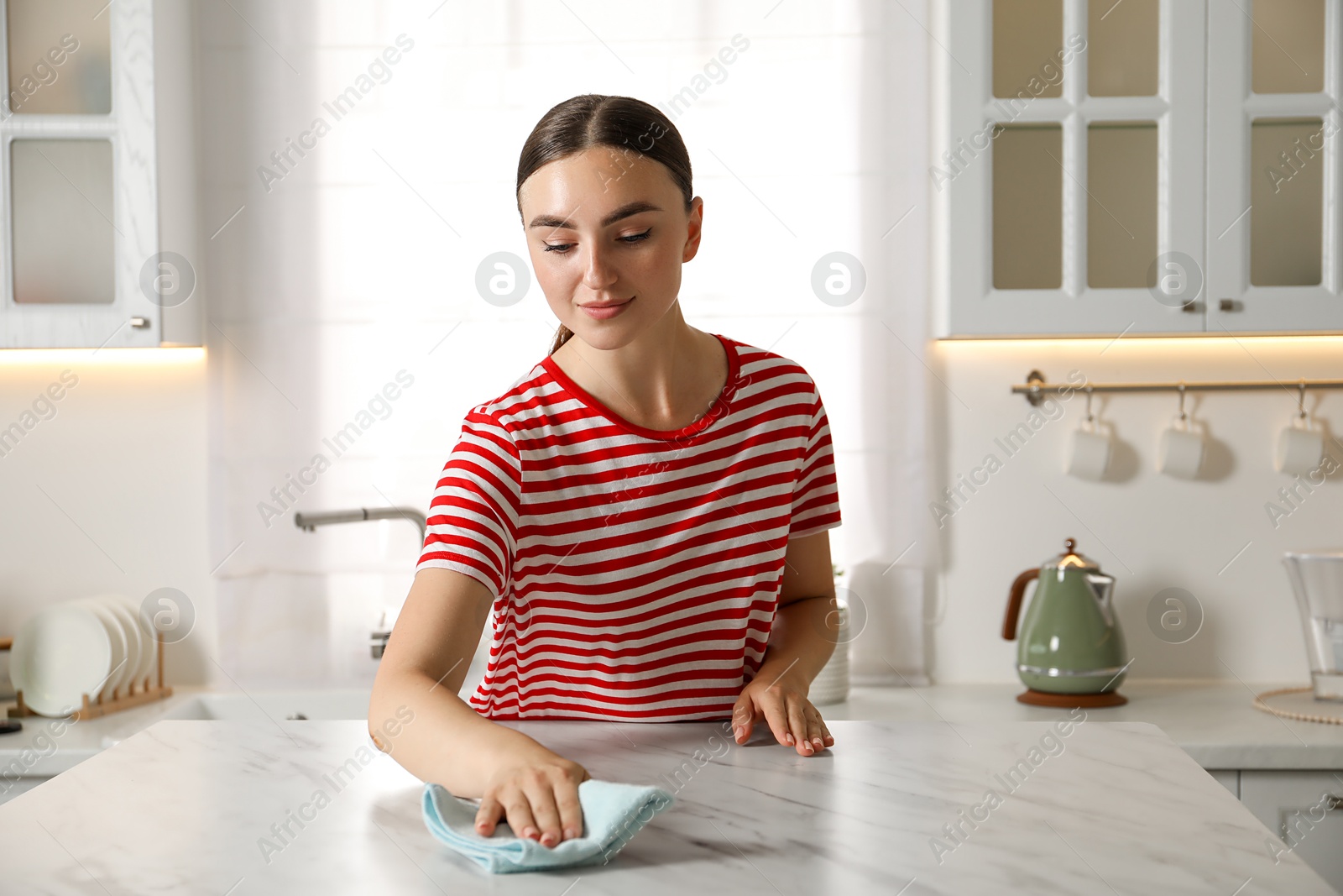 Photo of Beautiful young woman cleaning white marble table with rag in kitchen