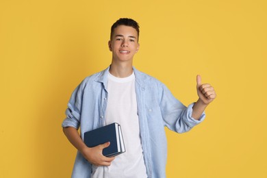 Portrait of teenage boy with books showing thumbs up on yellow background