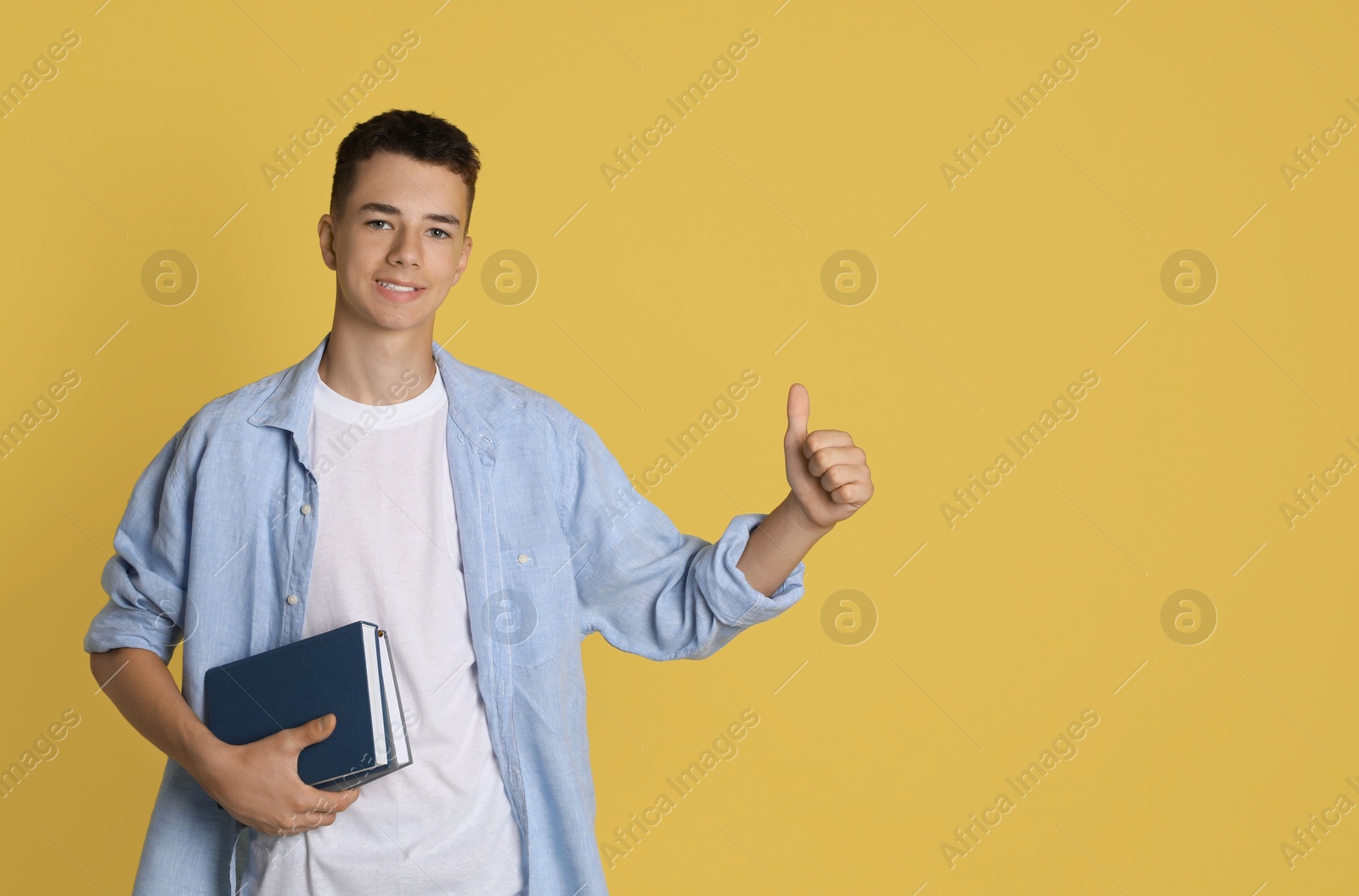 Photo of Portrait of teenage boy with books showing thumbs up on yellow background, space for text