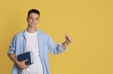 Photo of Portrait of teenage boy with books showing thumbs up on yellow background, space for text