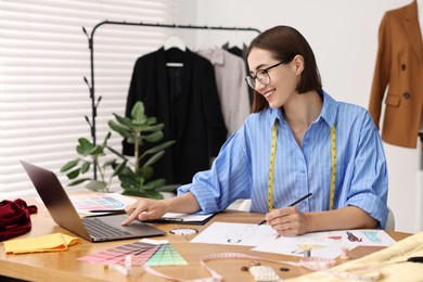 Fashion designer using laptop while drawing sketch of beautiful outfit at wooden table in workshop