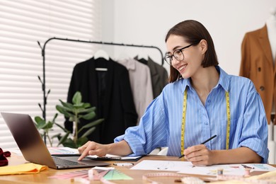 Photo of Fashion designer using laptop while drawing sketch of beautiful outfit at wooden table in workshop