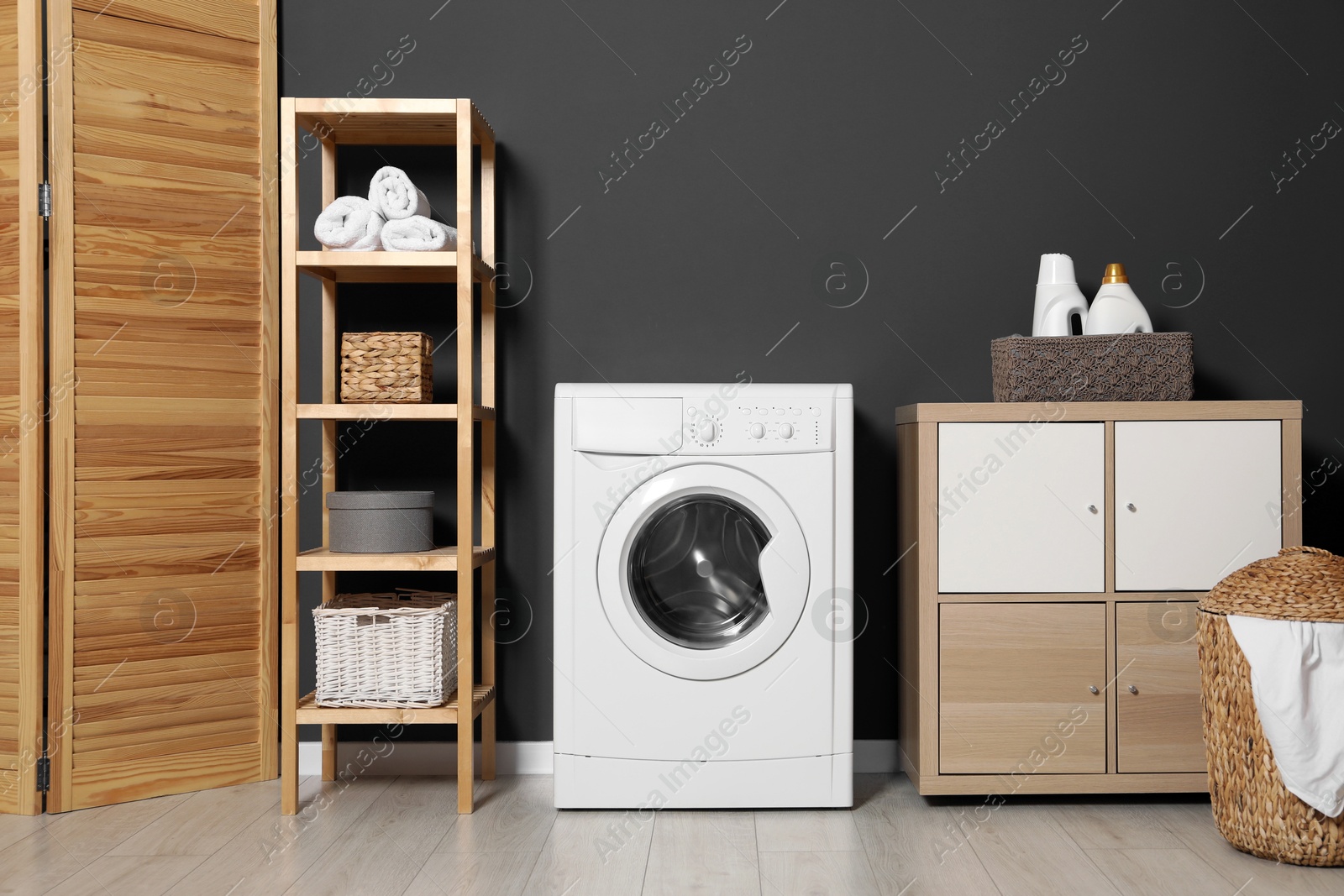 Photo of Washing machine, detergents in basket, wooden rack and storage cabinet indoors