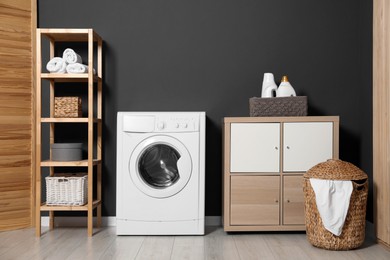 Photo of Washing machine, detergents in basket, wooden rack and storage cabinet indoors