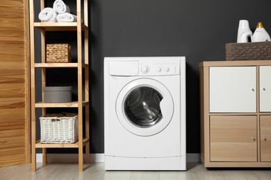 Photo of Washing machine, detergents in basket, wooden rack and storage cabinet indoors