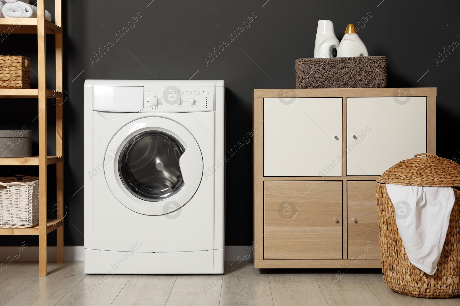 Photo of Washing machine, detergents in basket, wooden rack and storage cabinet indoors