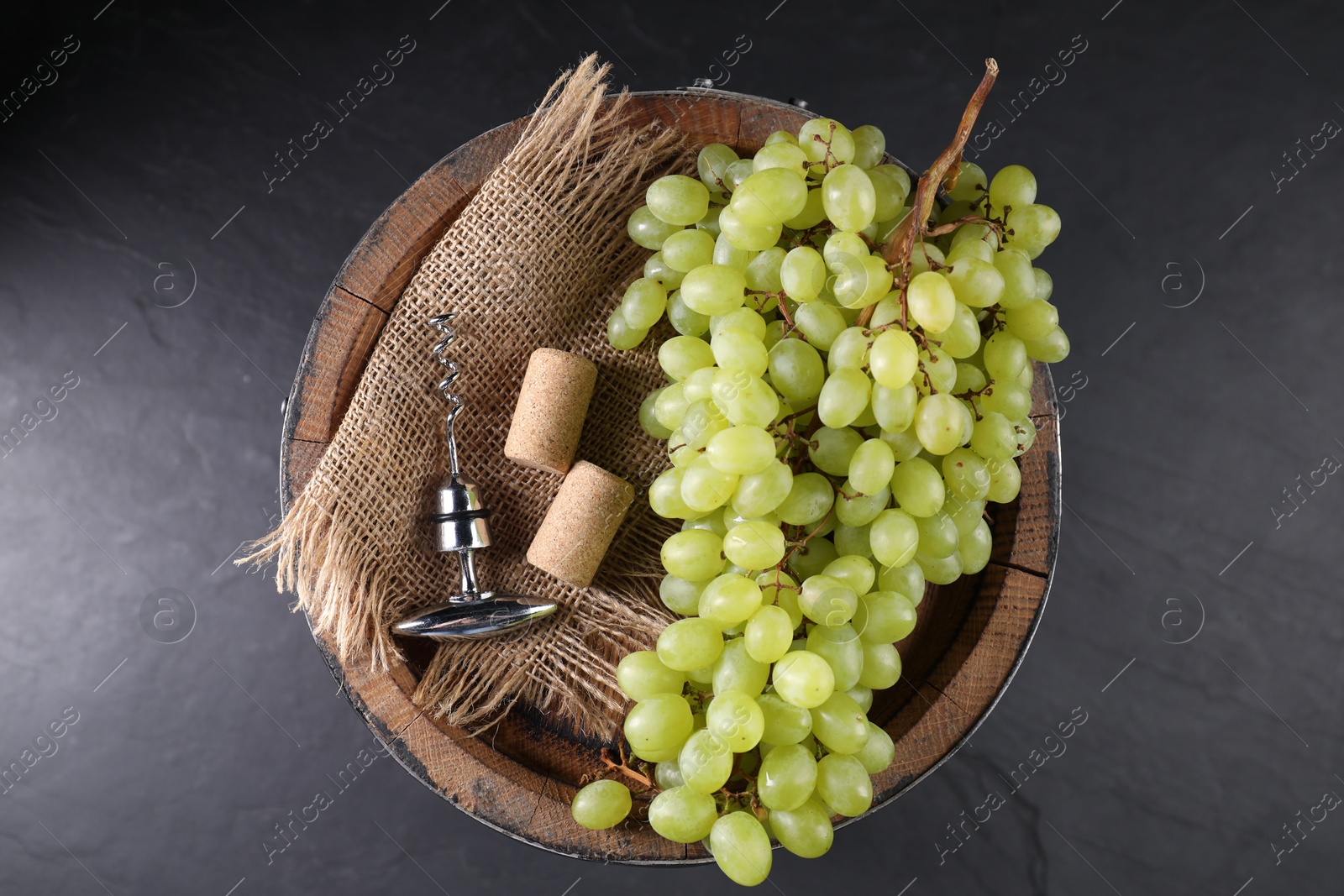 Photo of Corkscrew with metal handle, corks, grapes and wooden barrel on grey table, top view