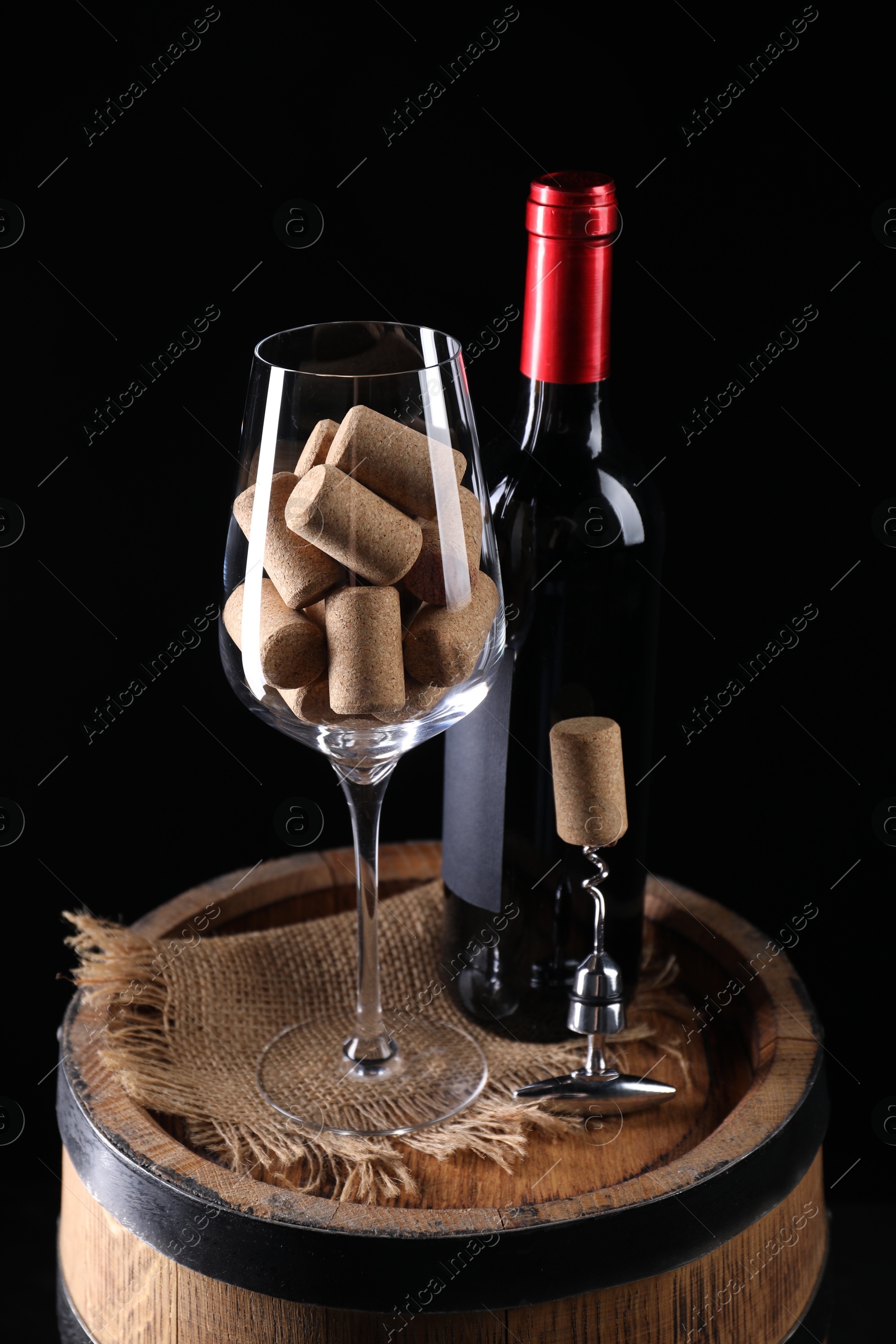 Photo of Corkscrew with metal handle, bottle of wine, glass and corks on wooden barrel against black background