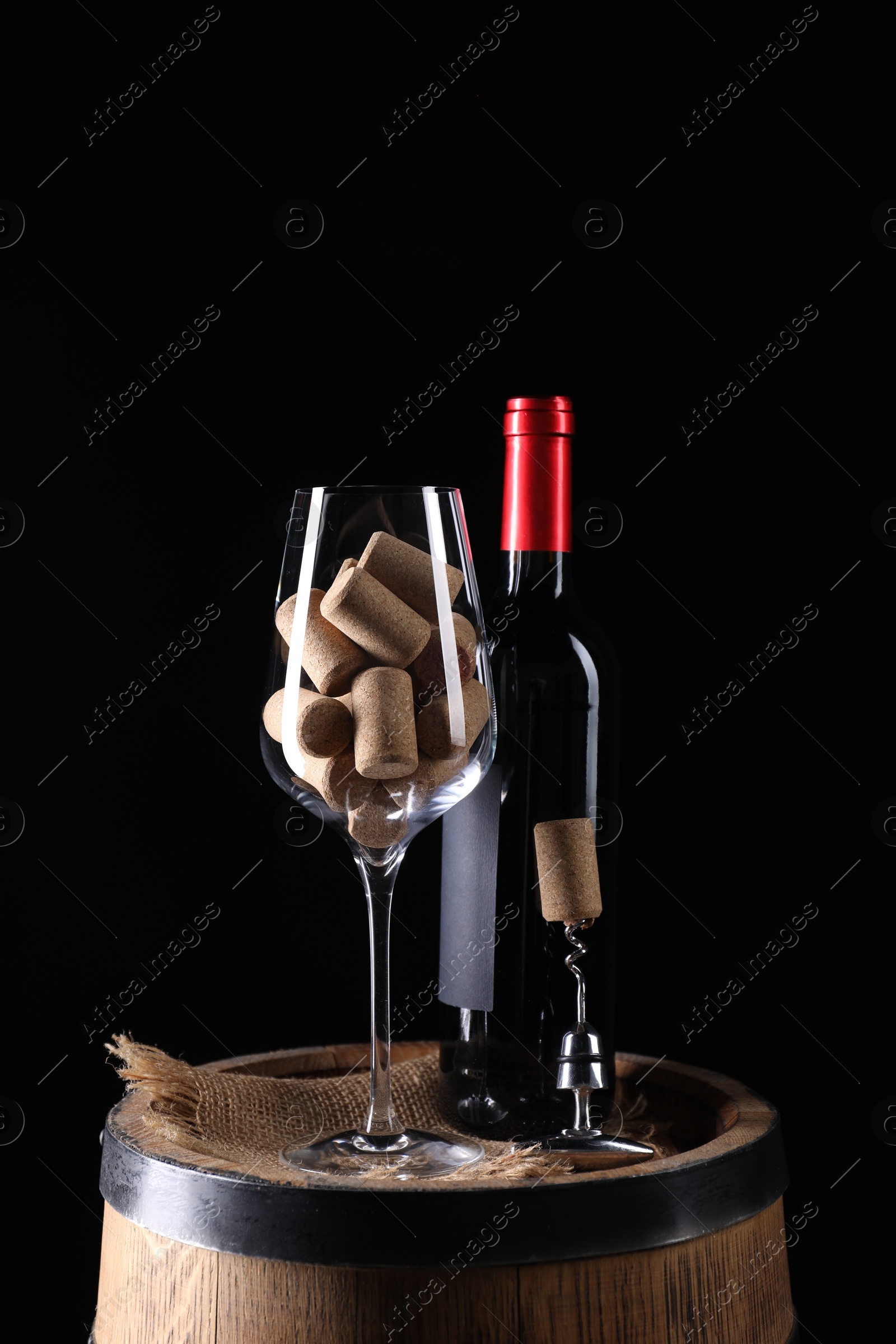 Photo of Corkscrew with metal handle, bottle of wine, glass and corks on wooden barrel against black background