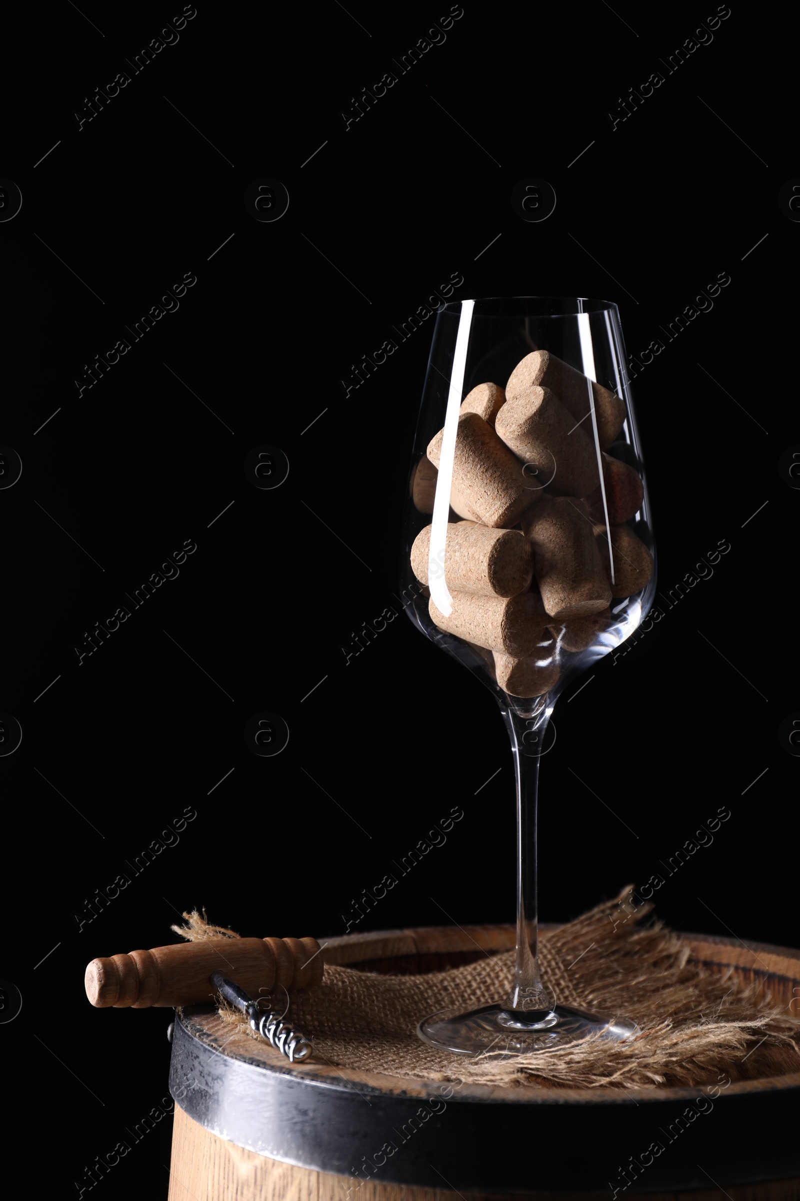 Photo of Corkscrew, glass and corks on wooden barrel against black background