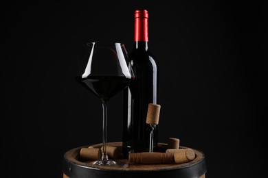 Photo of Corkscrew, bottle of wine, glass and corks on wooden barrel against black background