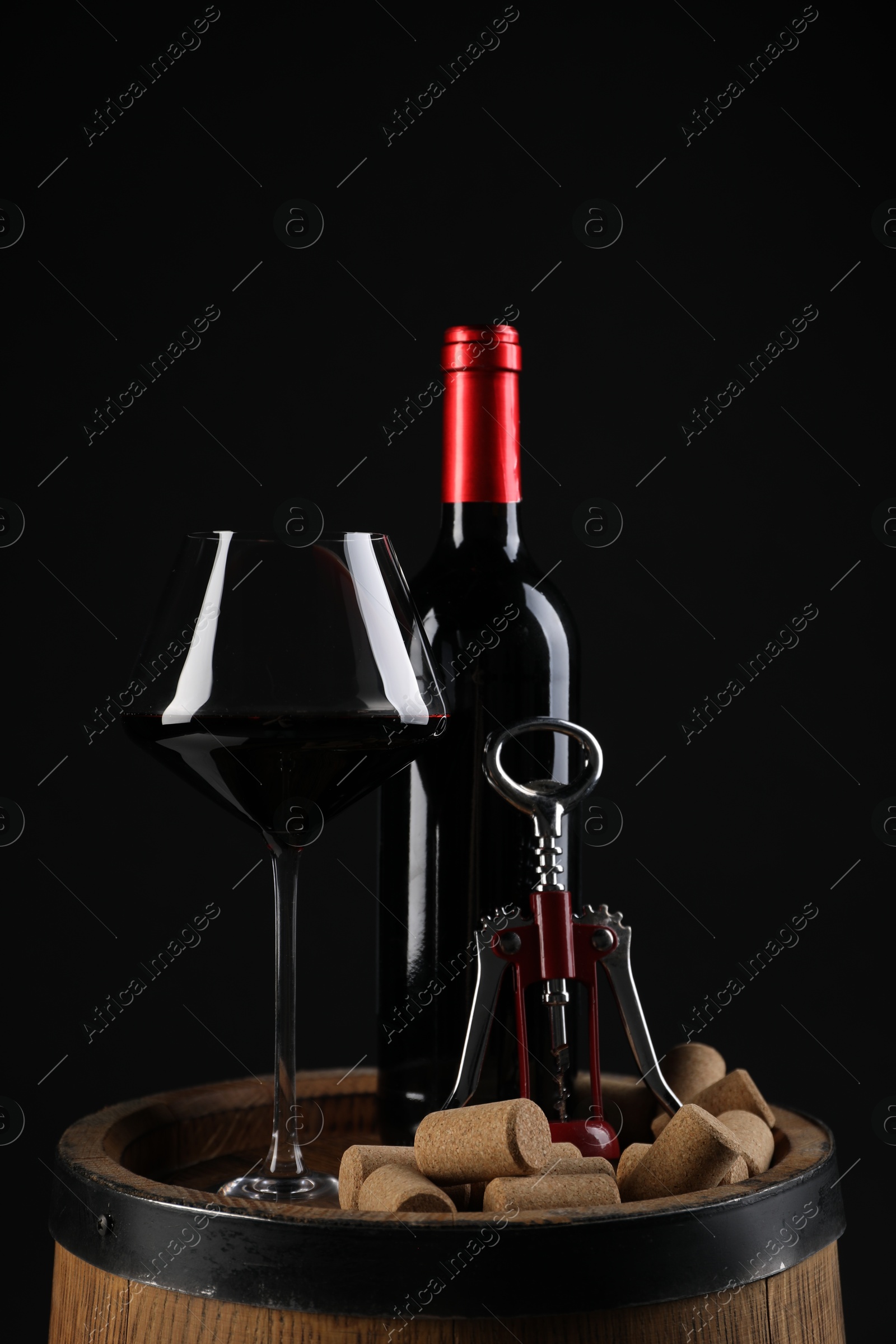 Photo of Wing corkscrew, bottle of wine, glass and corks on wooden barrel against black background