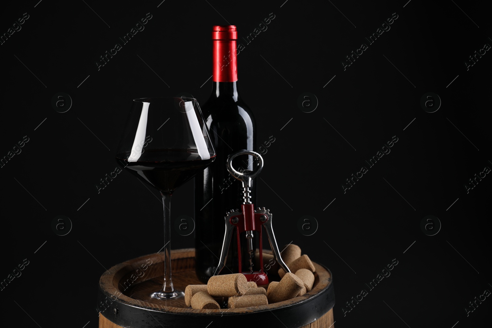 Photo of Wing corkscrew, bottle of wine, glass and corks on wooden barrel against black background