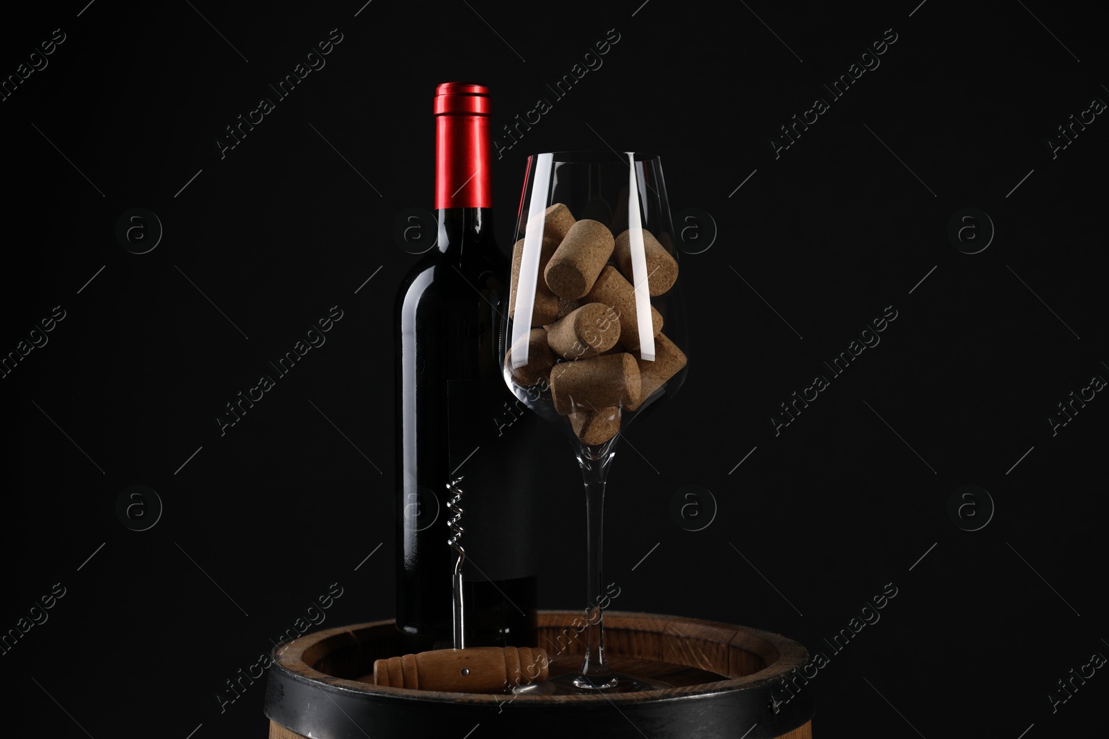 Photo of Corkscrew, bottle of wine, glass and corks on wooden barrel against black background