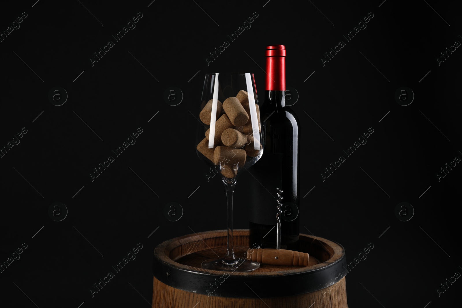 Photo of Corkscrew, bottle of wine, glass and corks on wooden barrel against black background