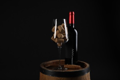 Photo of Corkscrew, bottle of wine, glass and corks on wooden barrel against black background