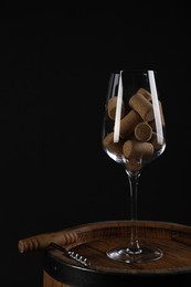 Photo of Corkscrew, glass and corks on wooden barrel against black background