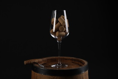 Photo of Corkscrew, glass and corks on wooden barrel against black background