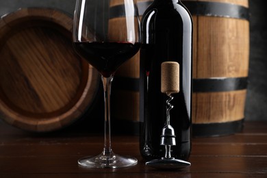 Photo of Corkscrew with metal handle, cork, bottle of wine, glass and barrels on wooden table, closeup