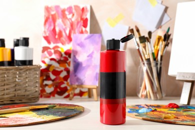 Red acrylic paint in bottle, small easels, palettes and brushes on wooden table indoors