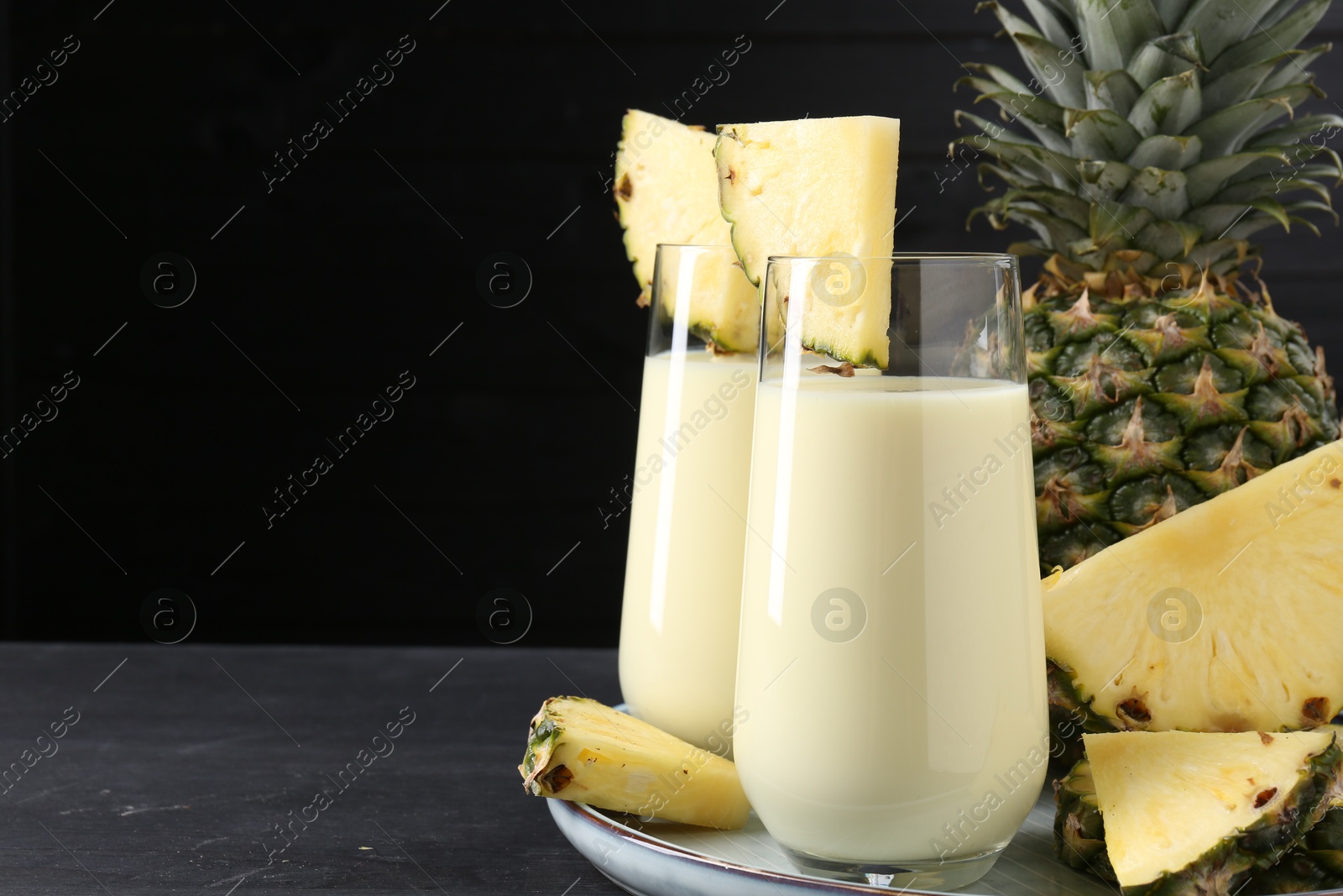 Photo of Tasty pineapple smoothie in glasses and fruit on black wooden table, closeup. Space for text