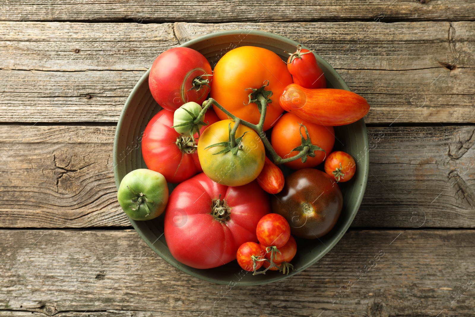 Photo of Different ripe tomatoes in bowl on wooden table, top view