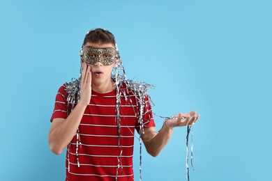 Surprised young man wearing carnival mask on light blue background. Space for text