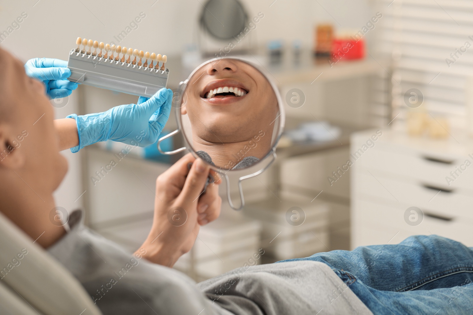 Photo of Patient looking in mirror while doctor holding teeth color chart indoors, closeup. Dental veneers