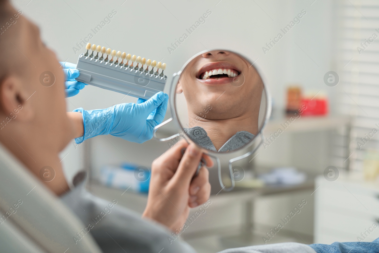 Photo of Patient looking in mirror while doctor holding teeth color chart indoors, closeup. Dental veneers