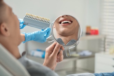 Patient looking in mirror while doctor holding teeth color chart indoors, closeup. Dental veneers