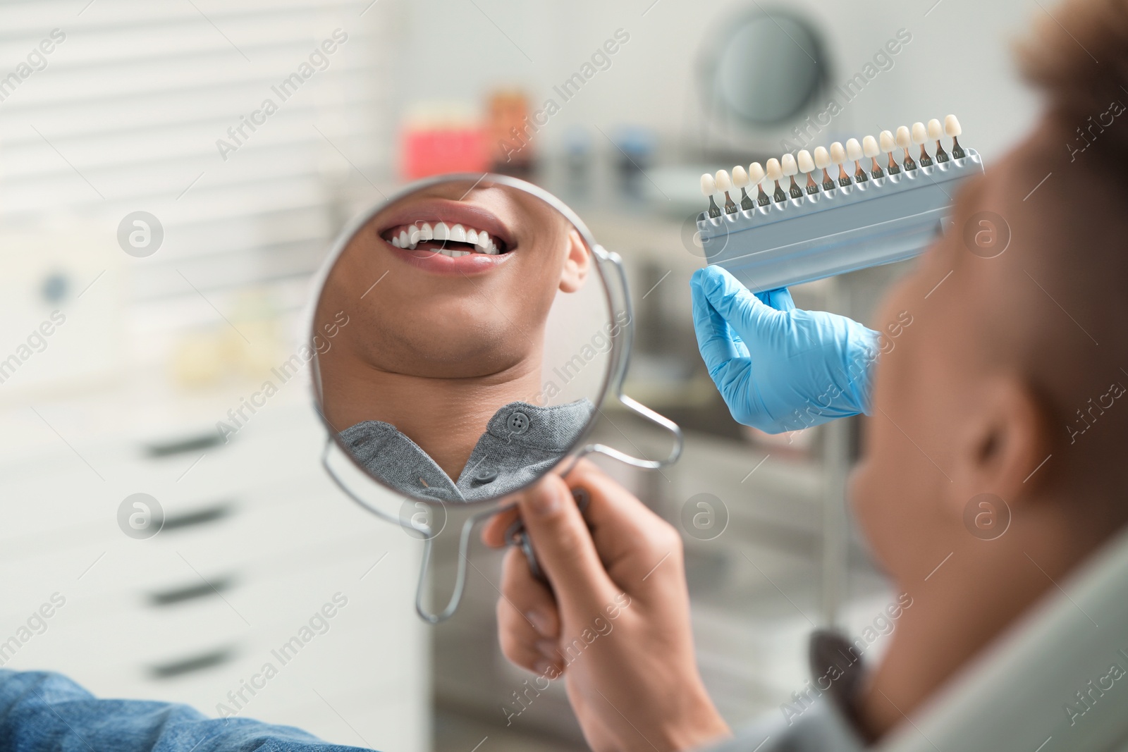 Photo of Patient looking in mirror while doctor holding teeth color chart indoors, closeup. Dental veneers