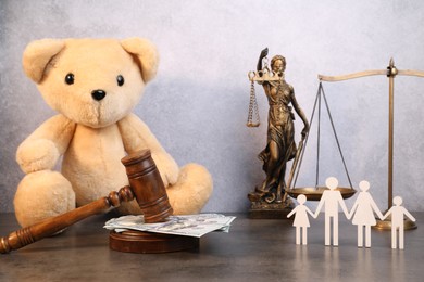 Photo of Alimony. Toy bear, wooden gavel with dollar banknotes, scales, figures of family and statue of Lady Justice on grey table