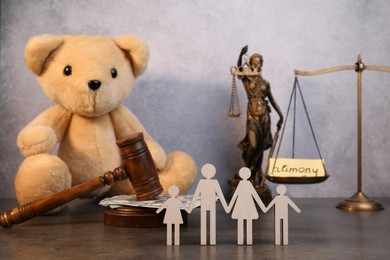 Photo of Alimony. Toy bear, wooden gavel with dollar banknotes, scales, figures of family and statue of Lady Justice on grey table