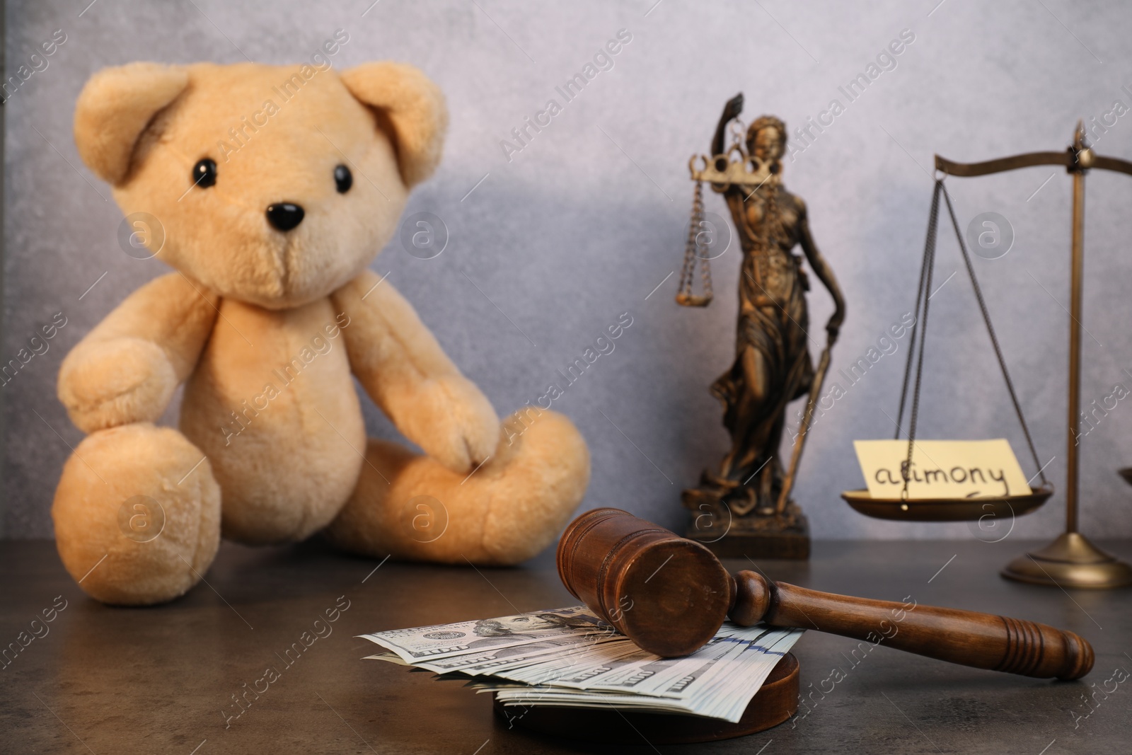 Photo of Alimony. Toy bear, wooden gavel with dollar banknotes, scales and statue of Lady Justice on grey table