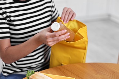 Photo of Woman putting bottle of drink into school lunch bag at wooden table indoors, closeup