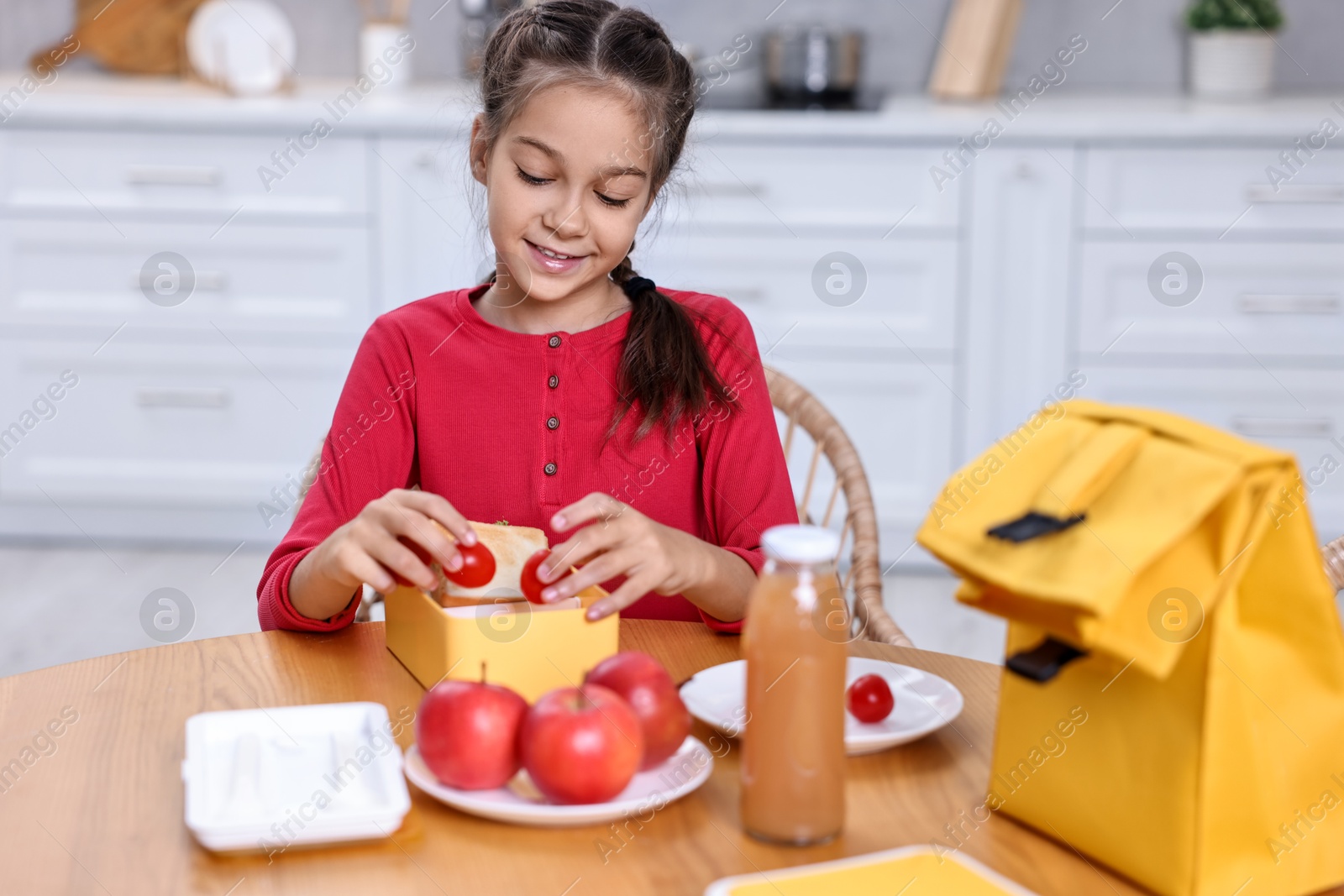 Photo of Cute girl putting tomatoes into lunch box at wooden table in kitchen. Preparing for school