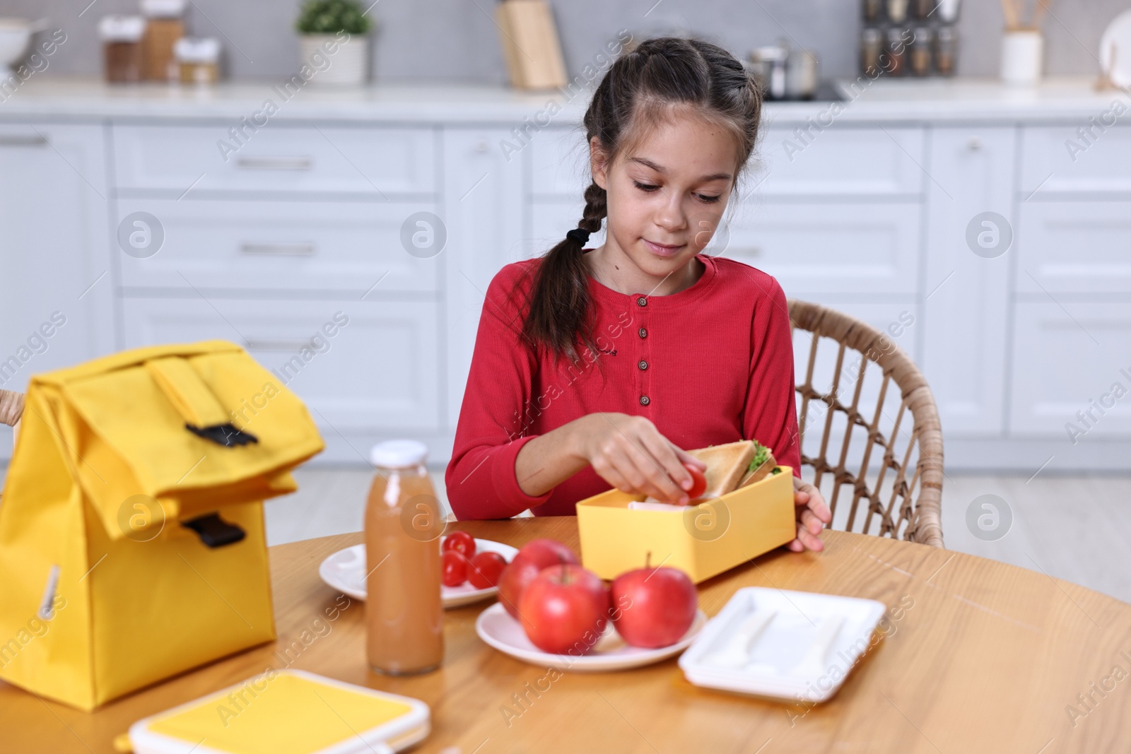 Photo of Cute girl putting tomatoes into lunch box at wooden table in kitchen. Preparing for school