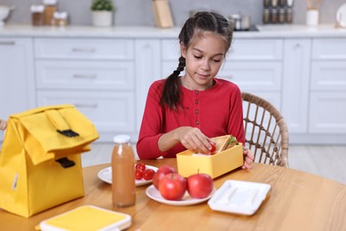 Cute girl putting tomatoes into lunch box at wooden table in kitchen. Preparing for school