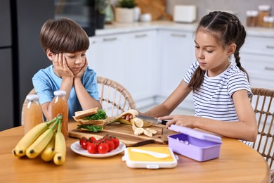 Photo of Cute children with healthy snacks preparing lunch box at wooden table in kitchen