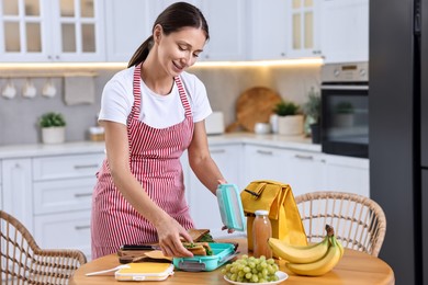 Smiling mother packing school lunch box with healthy snacks at wooden table in kitchen