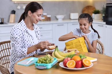 Cute girl and her mother preparing lunch box with healthy snacks at wooden table in kitchen