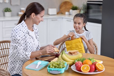 Photo of Cute girl and her mother preparing lunch box with healthy snacks at wooden table in kitchen