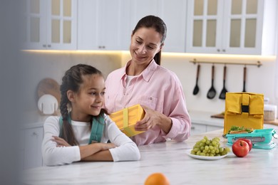 Photo of Smiling mother putting lunch box into daughter`s backpack in kitchen. Preparing for school