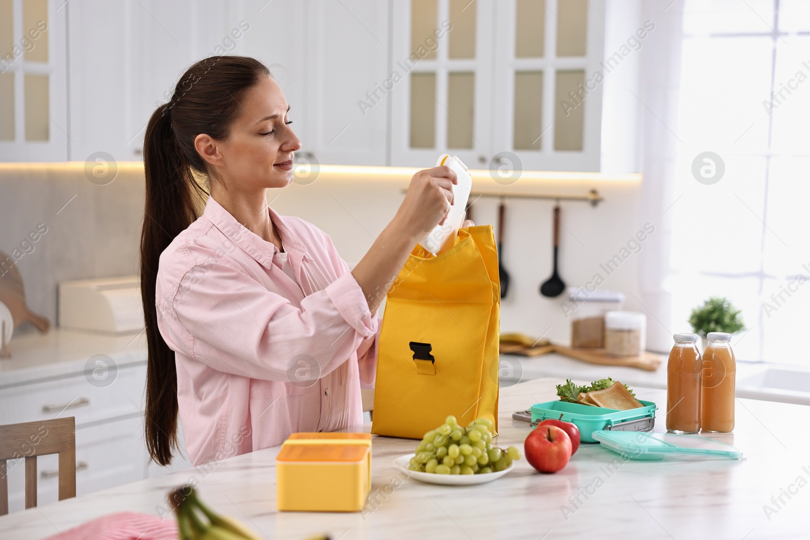 Photo of Woman with bag packing school lunch box and healthy food at white marble table in kitchen