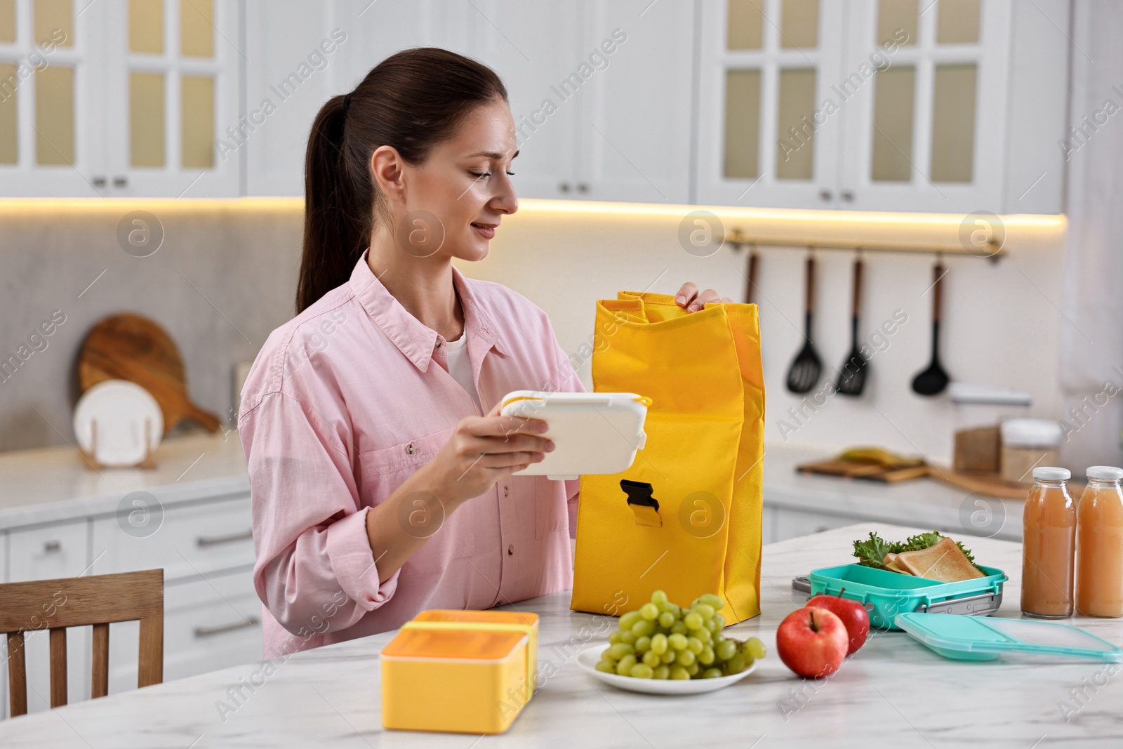 Photo of Woman with bag packing school lunch box and healthy food at white marble table in kitchen