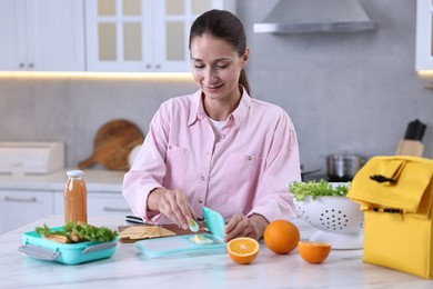 Photo of Smiling woman making snacks for school lunch box at white marble table in kitchen